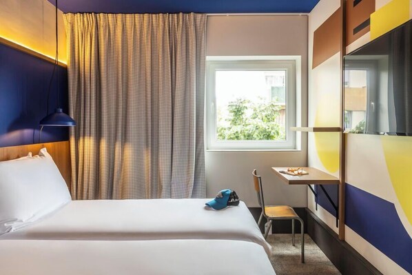 A hotel room with a bed, a desk, a chair, a window with curtains, and a view of greenery outside. - greet hotel Marseille Parc Chanot Vélodrome by Accor (Marseille)