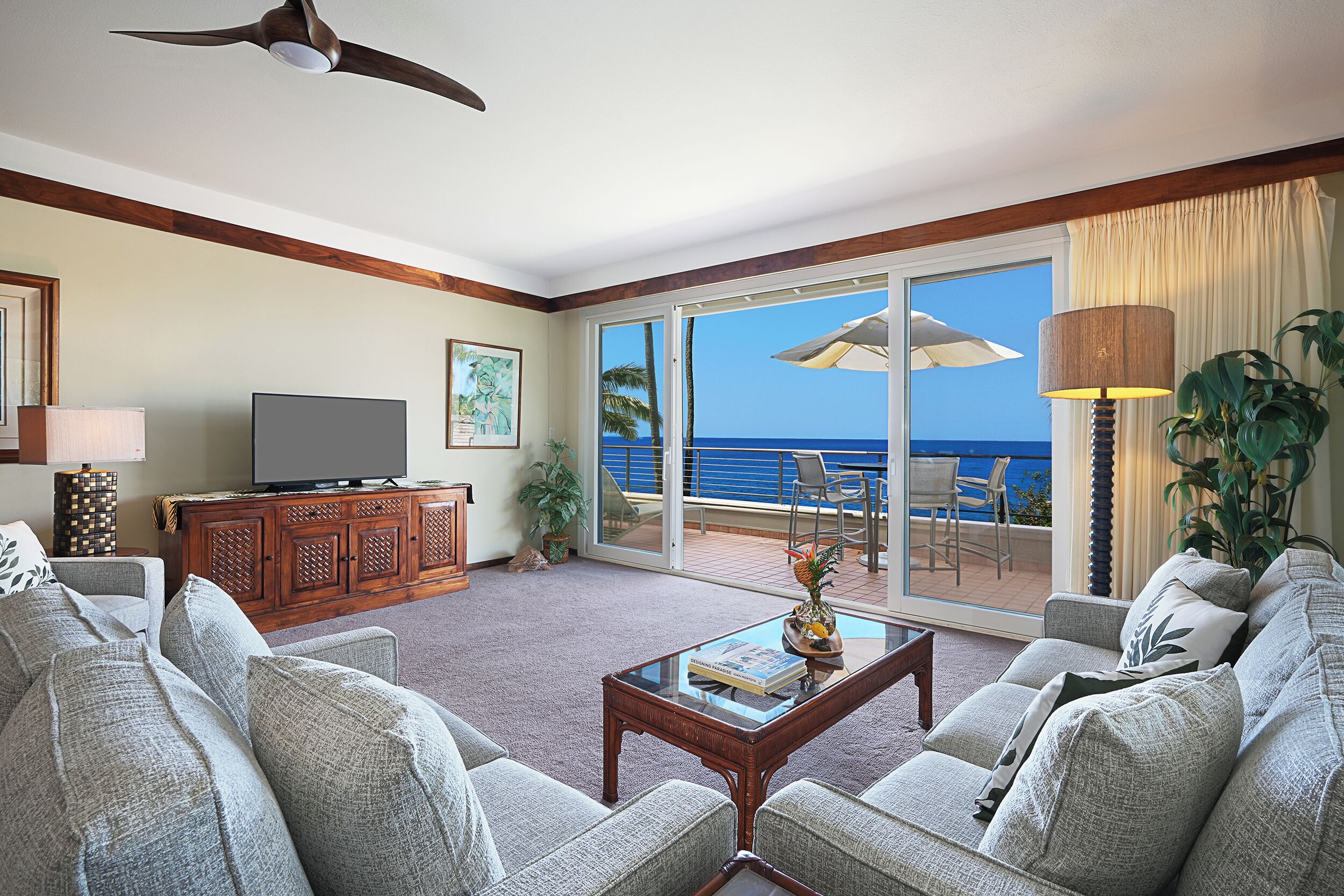 A living room with a television, a coffee table with a book, and a view of the ocean through large sliding glass doors.