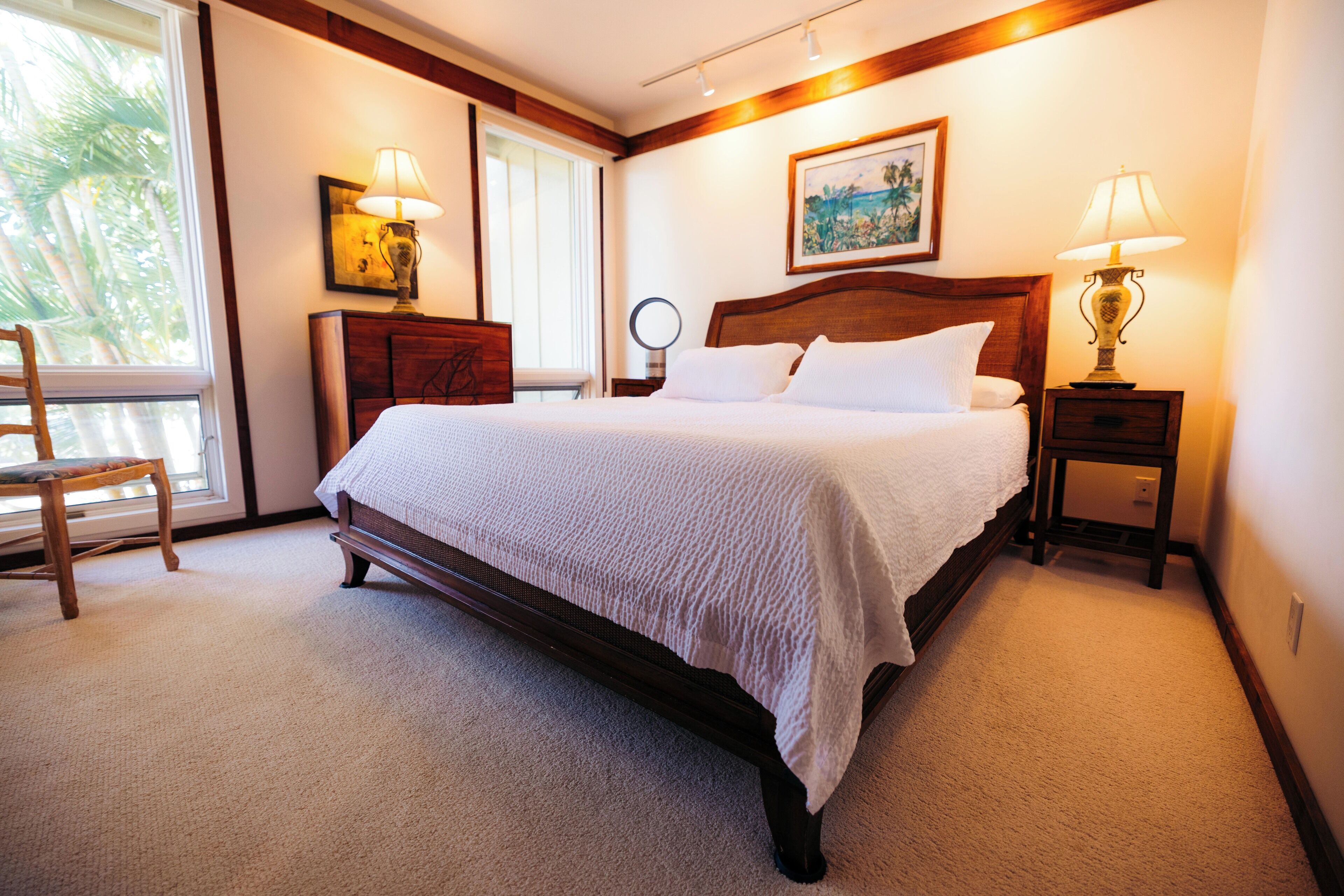Une chambre à coucher bien aménagée, avec un grand lit, des tables de chevet, une chaise et une fenêtre donnant sur des palmiers.