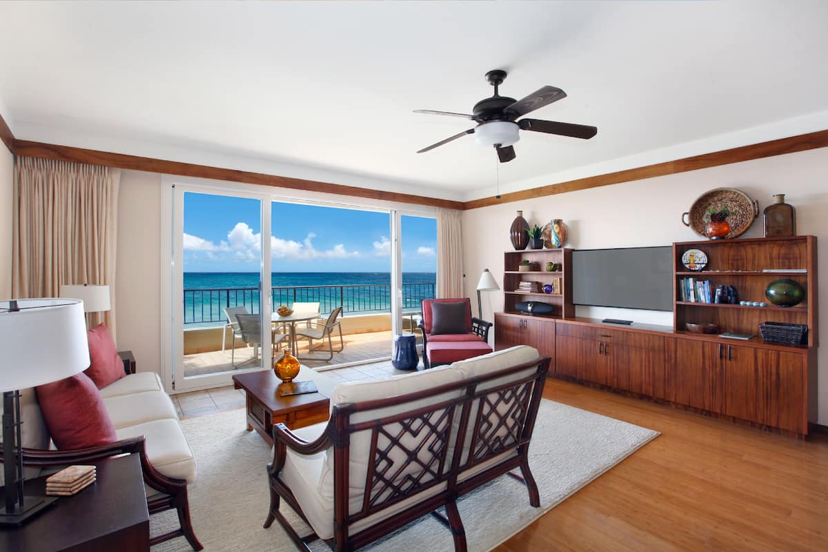 A spacious living room with a large TV, wooden furniture, and a balcony with a view of the ocean.