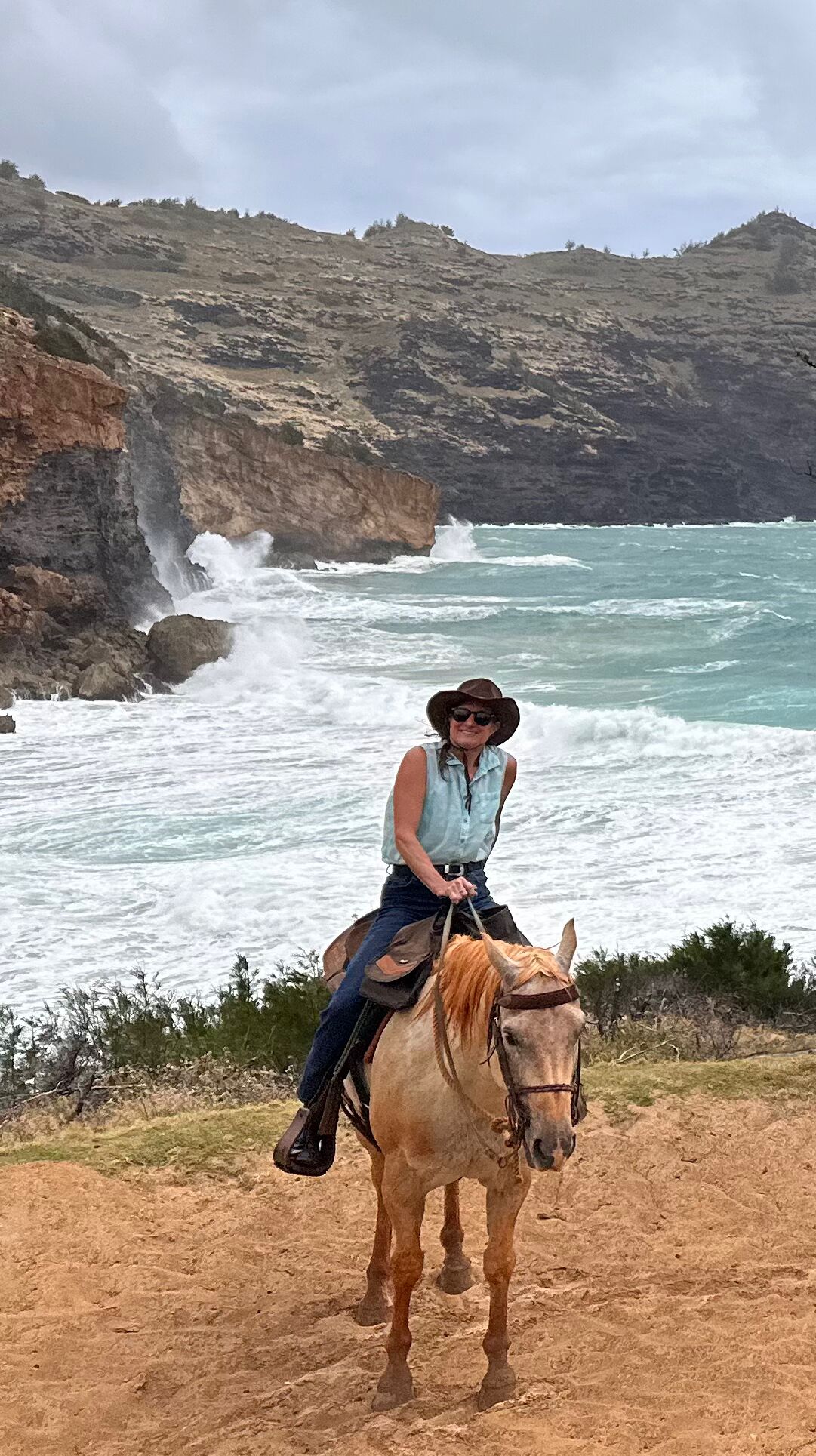 A person riding a horse on a rocky coastal path with ocean waves crashing against the cliffs in the background.