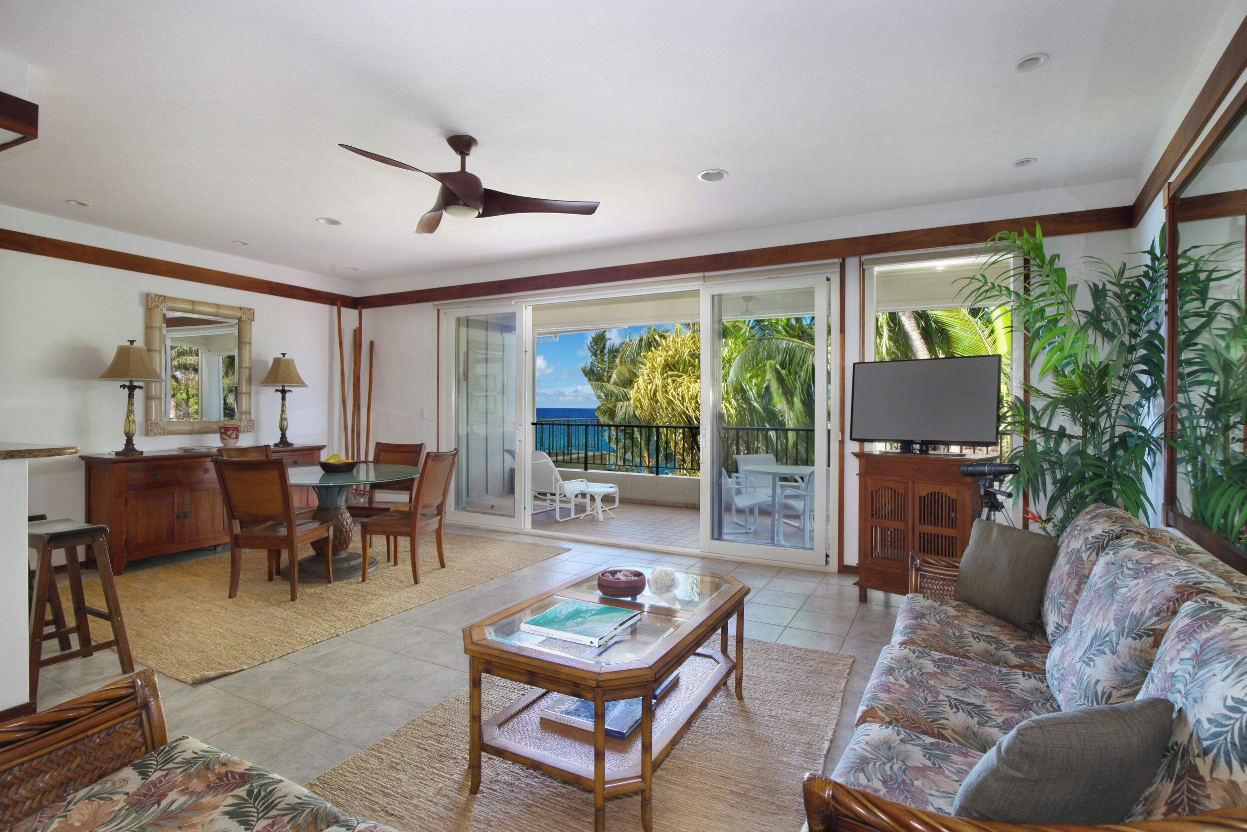 A spacious living room with a large sofa, a coffee table, and a TV. There is a dining area with chairs and a table, and a balcony with a view of the ocean.