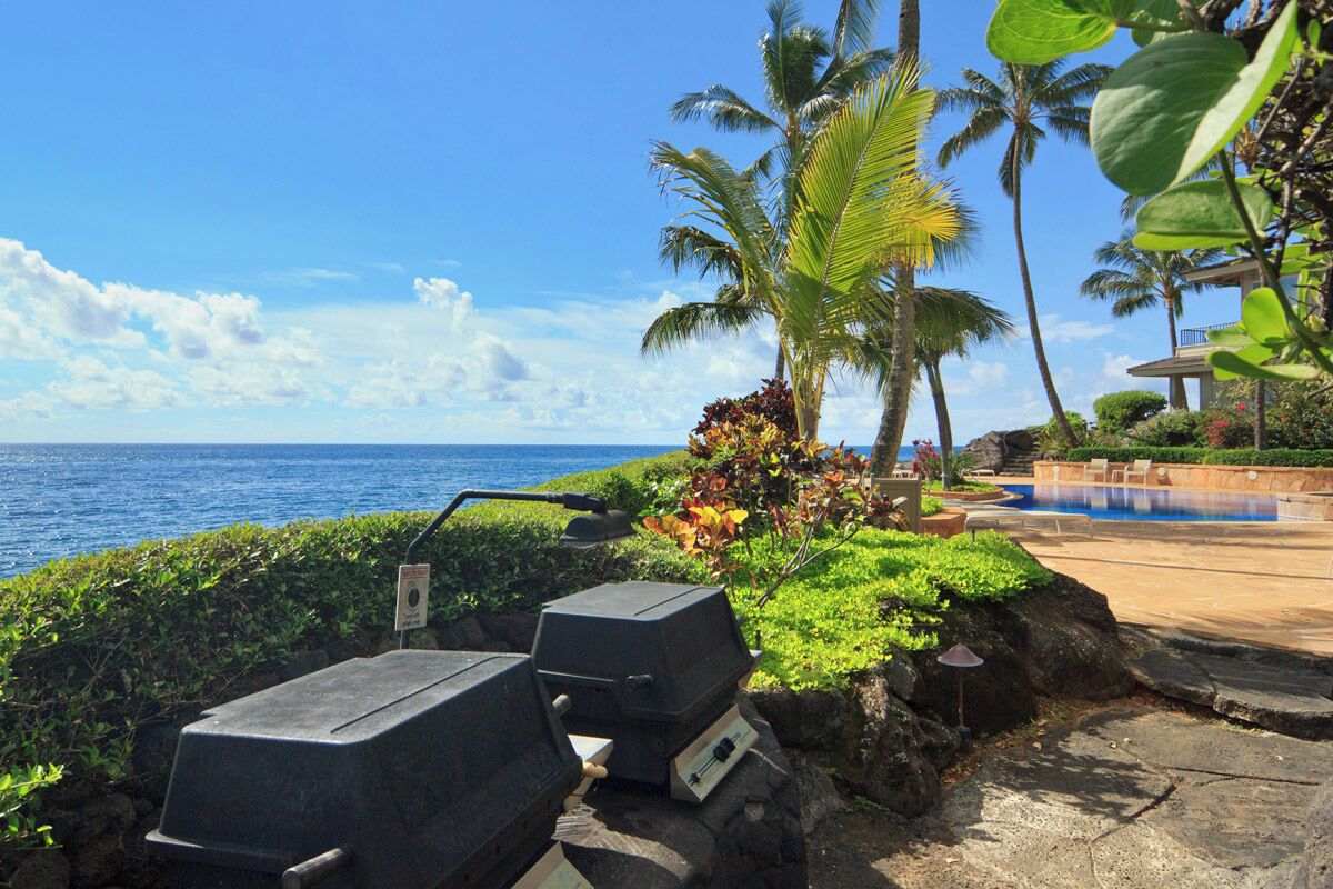 Un espace aménagé au bord de la piscine, offrant une vue sur l’océan, les palmiers et un barbecue.