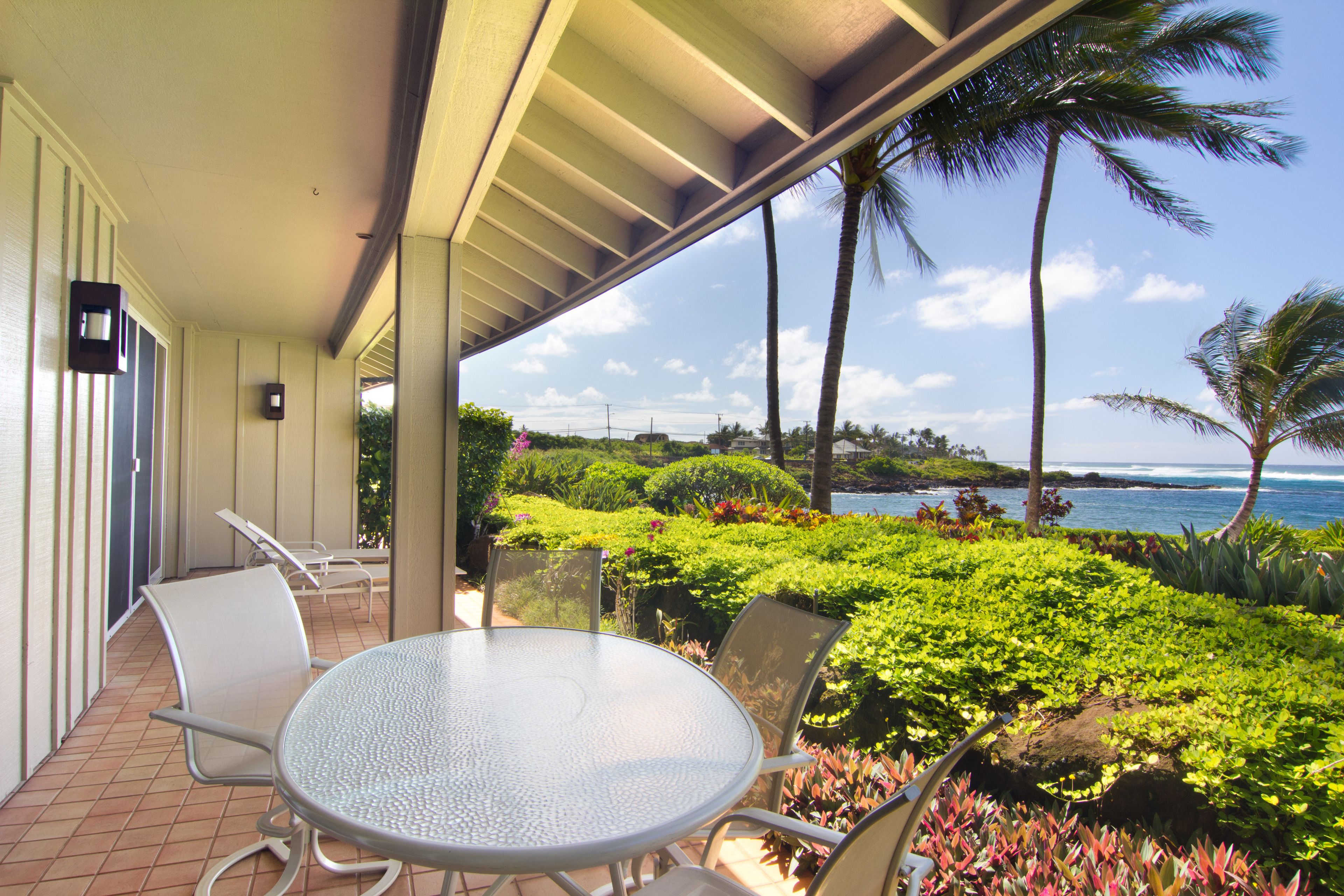 A covered patio with a round table and chairs, overlooking a lush garden and the ocean.