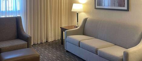 A hotel room with a sofa, armchair, and a lamp on a side table.