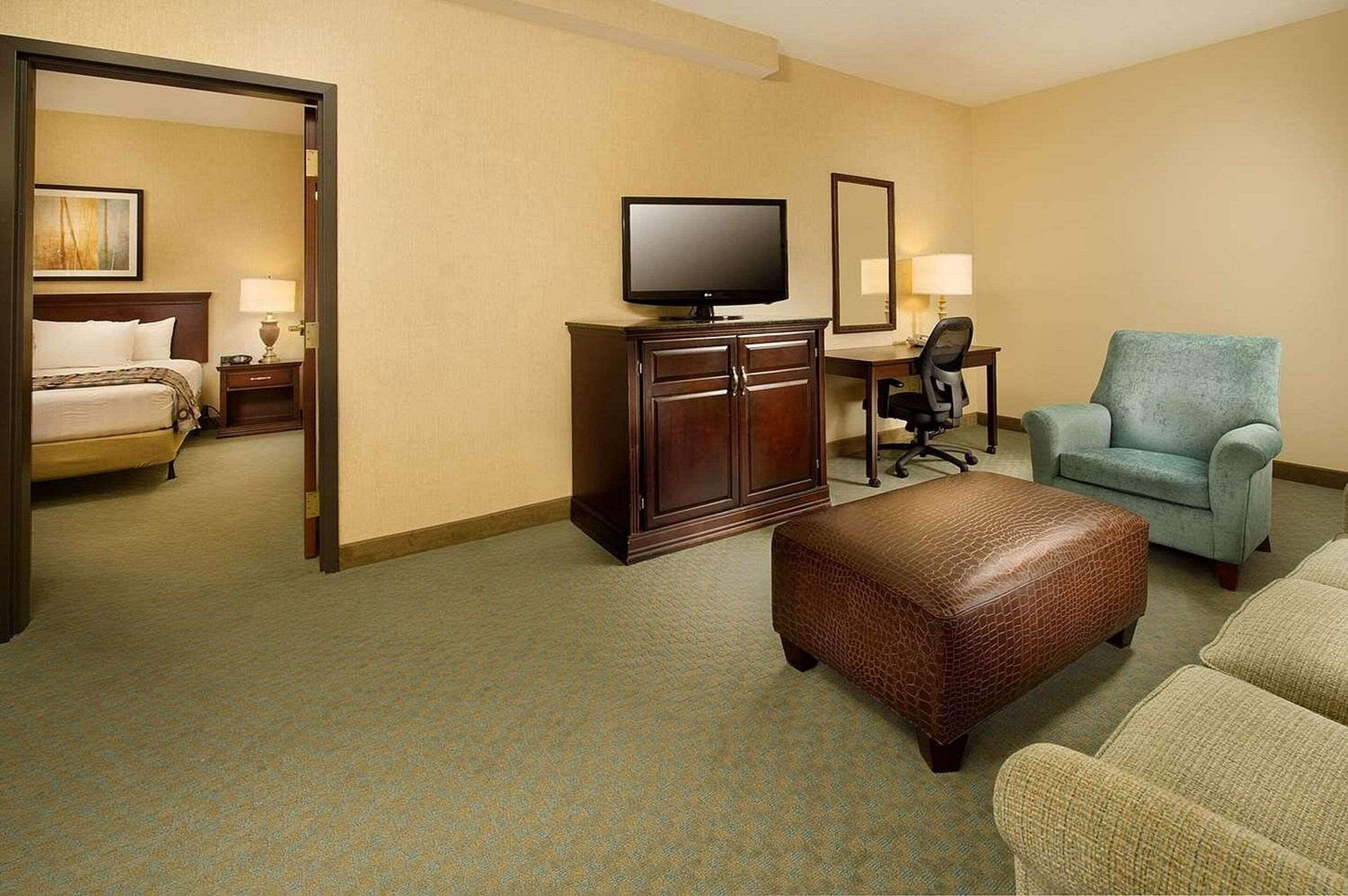 Quarto de hotel com cama, televisão, escrivaninha com cadeira, sofá e apoio para os pés.