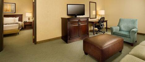 Quarto de hotel com cama, televisão, escrivaninha com cadeira, sofá e apoio para os pés.
