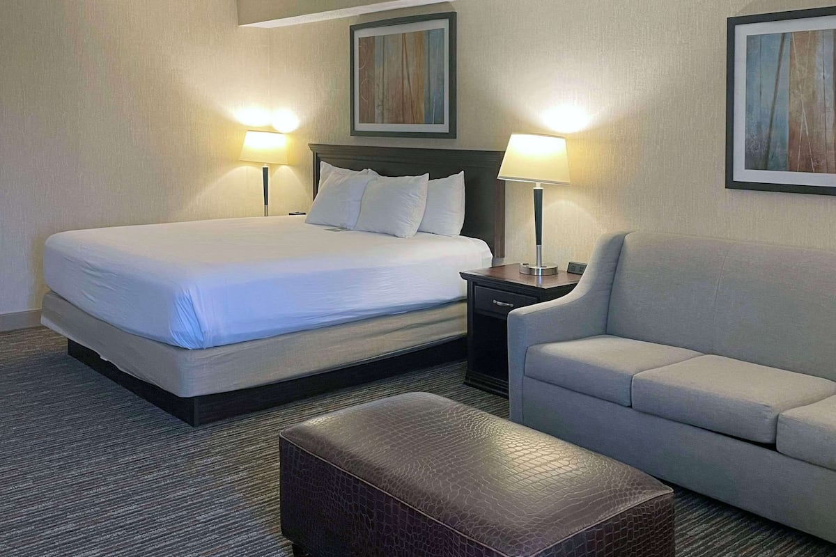 A hotel room with a large bed, two bedside lamps, a grey sofa, and a small ottoman.