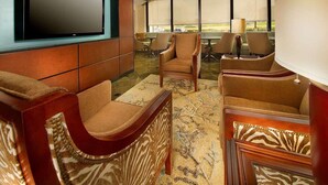 A hotel lobby with a large flat-screen TV mounted on a wall, several upholstered chairs, a patterned carpet, and large windows offering a view of the outdoors. - Prado Hotel (Ridgeland)
