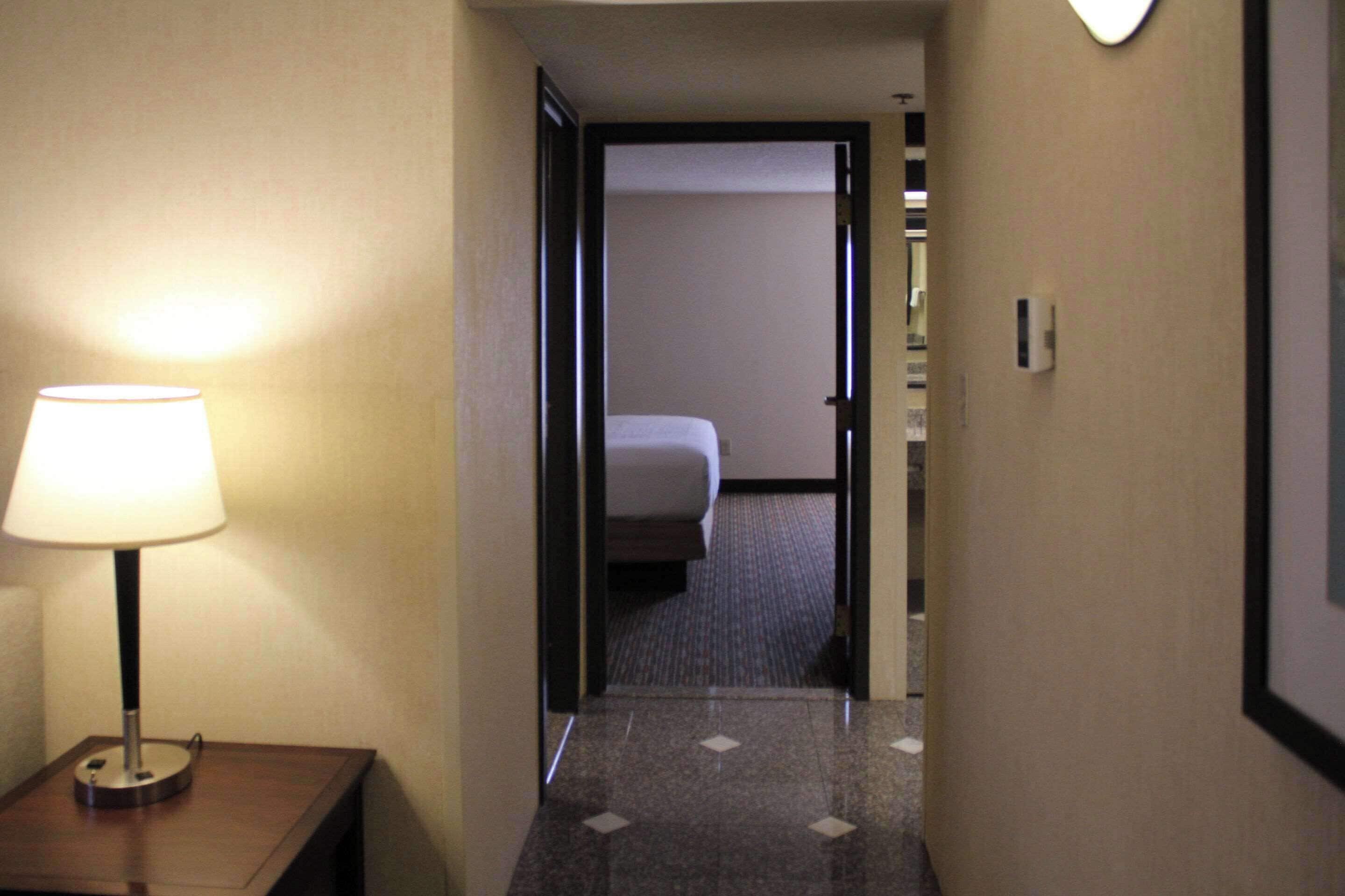 A hotel hallway with a bed in the background, a lamp on the left, and a door on the right.