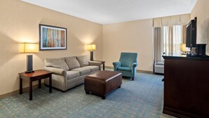 A hotel room with a sofa, a chair, a wooden table, a lamp, a television, and a framed picture on the wall. - Prado Hotel (Ridgeland)