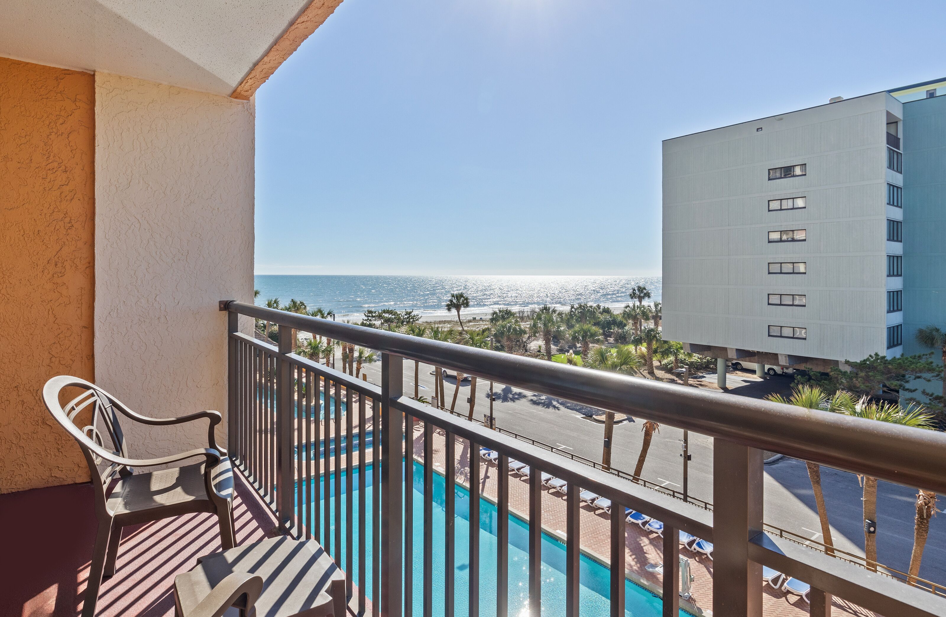 A balcony with a view of the ocean, a pool, and a building in the background.