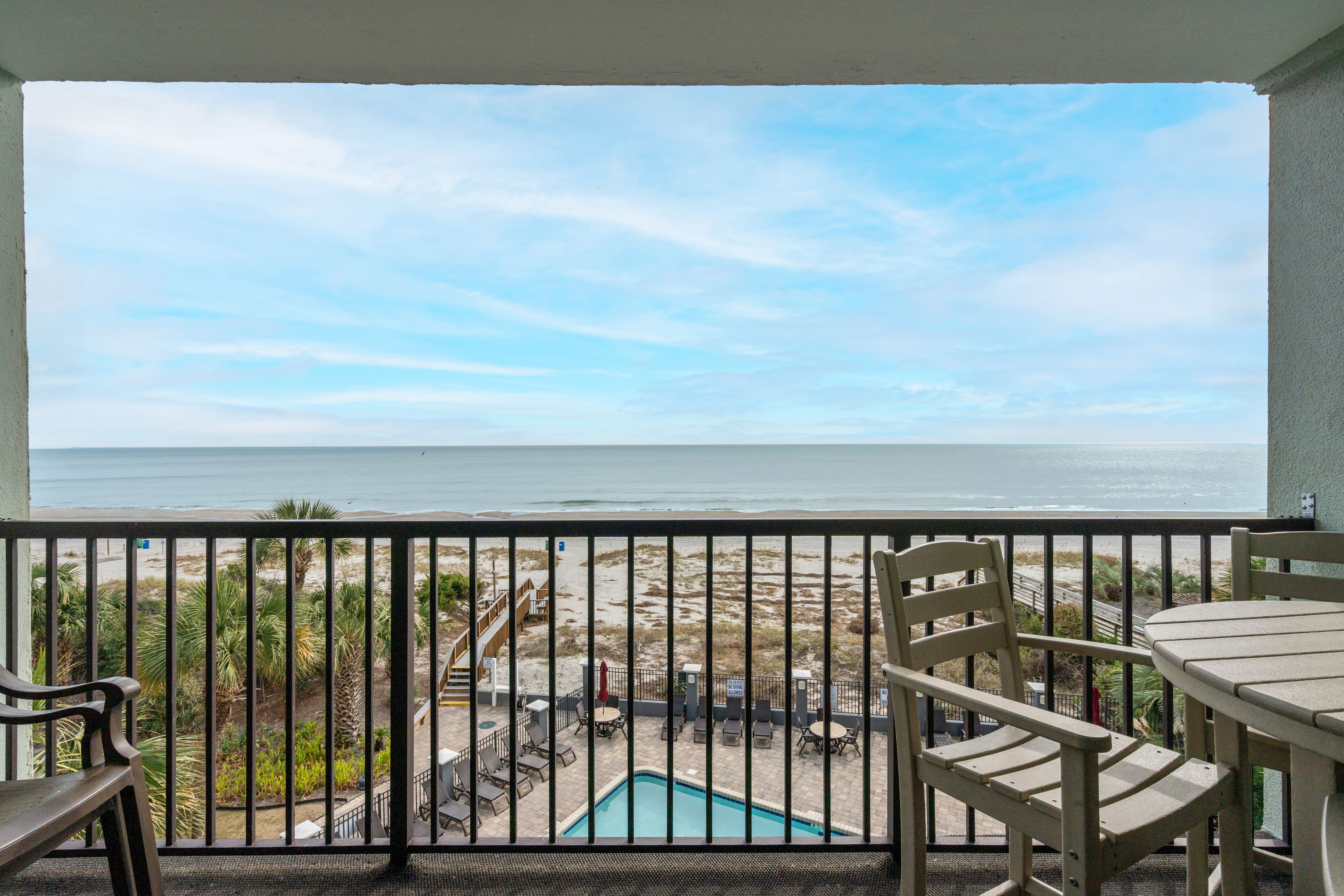 A balcony with a pool, chairs, and a view of the beach and ocean.