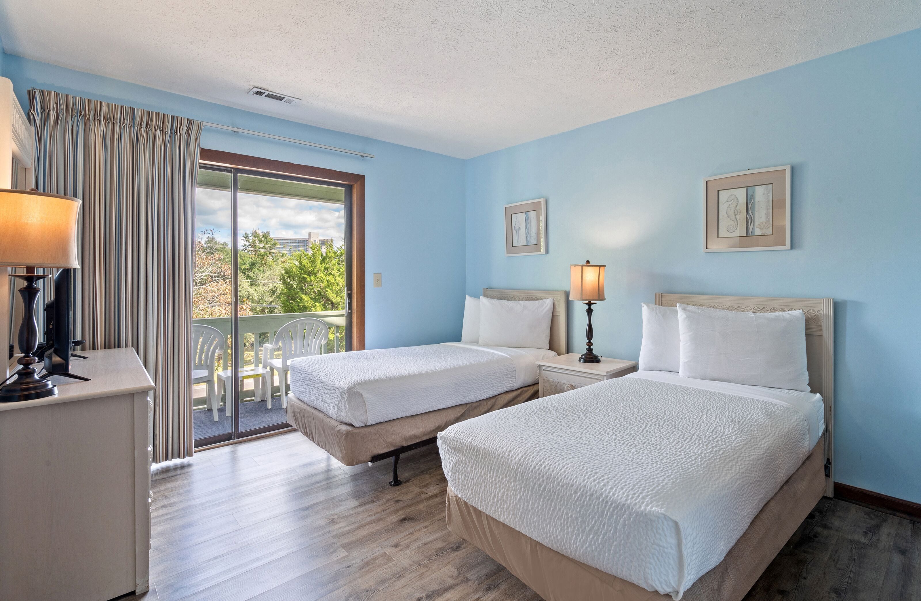 A hotel room with two beds, a desk, a chair, a lamp, and a view of the outdoors.