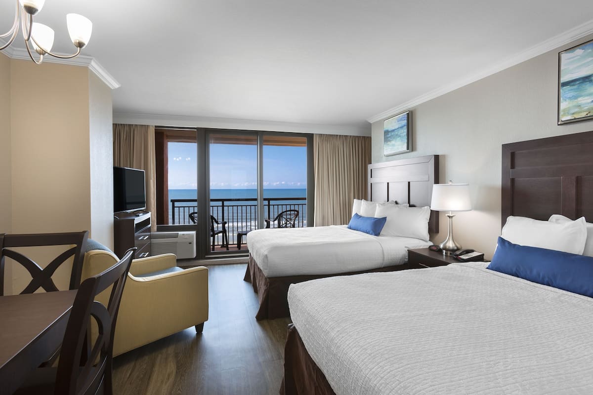 Habitación de hotel con dos camas, una mesa de comedor, sillas, televisor y balcón con vista al mar.
