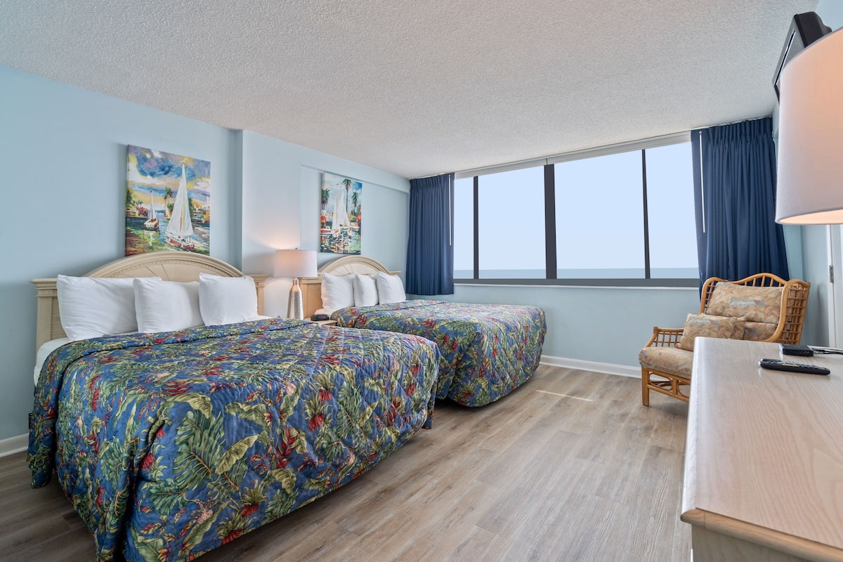 Habitación de hotel con dos camas, un escritorio de madera, una silla y un amplio ventanal con cortinas.