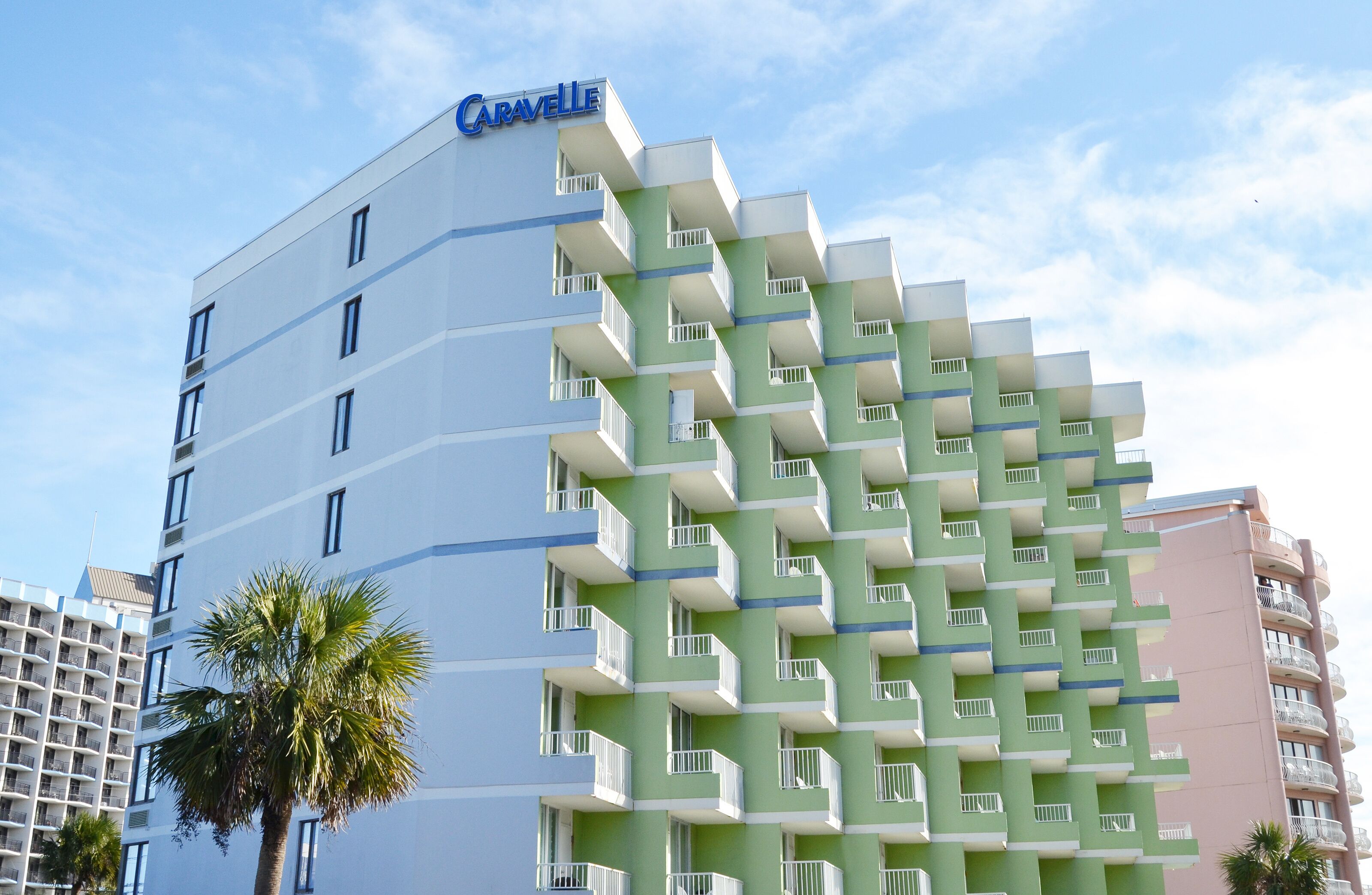 Ein mehrgeschossiges Hotel mit einer grün-weißen Fassade, Balkonen auf jeder Etage und einer Palme im Vordergrund.