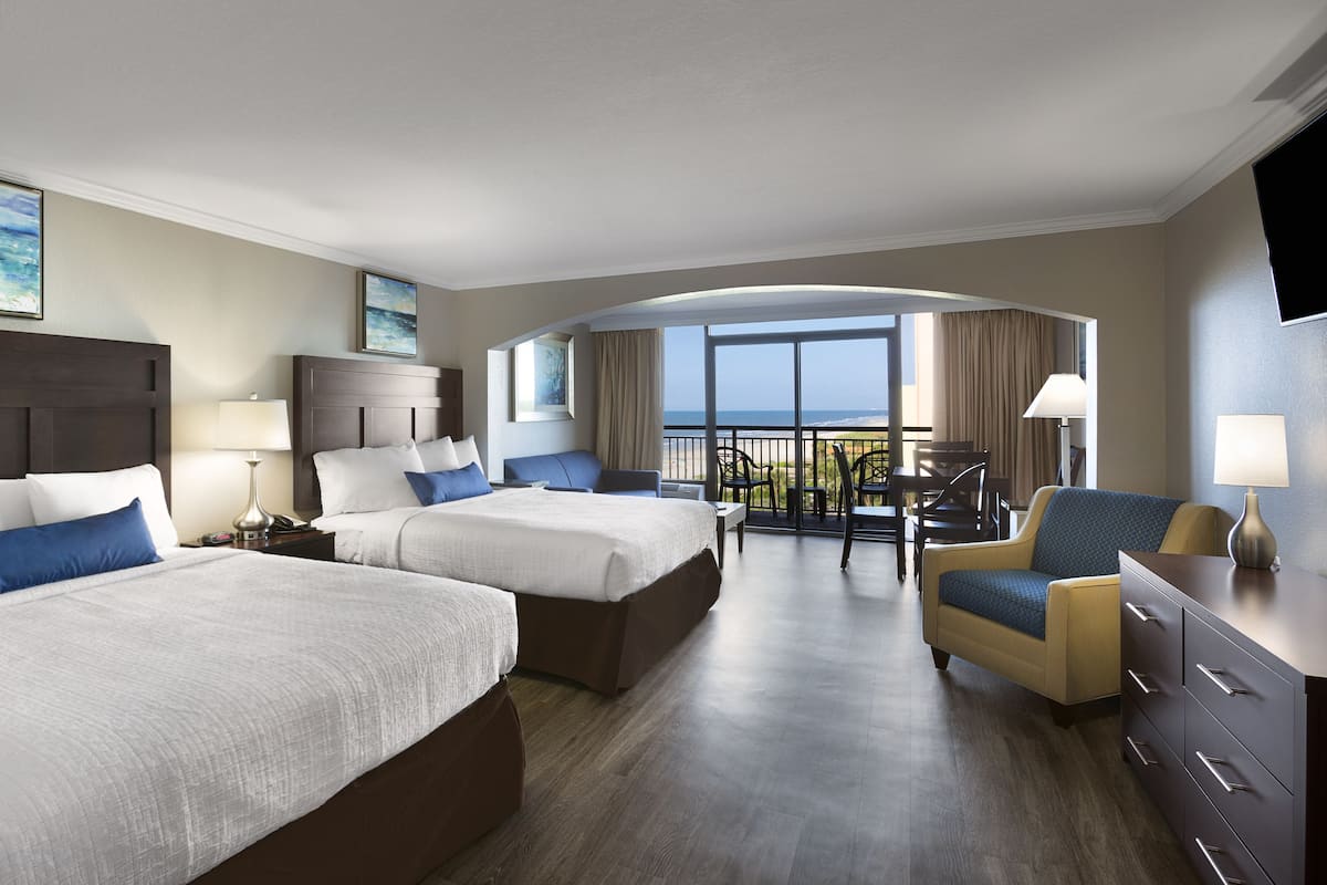 Una habitación de hotel con dos camas, una zona de estar con vista al océano y un balcón.