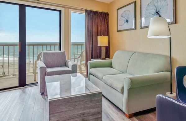 A hotel room with a balcony, a sofa, a chair, a table, and a lamp.