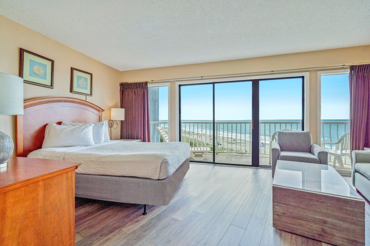 Habitación de hotel con una cama grande, un escritorio, una silla y vistas a la playa desde las puertas del balcón.