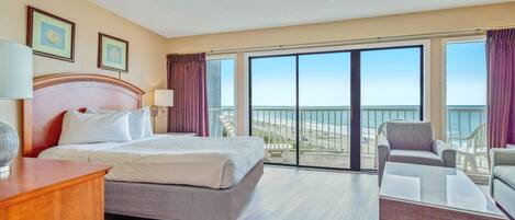 Habitación de hotel con una cama grande, un escritorio, una silla y vistas a la playa desde las puertas del balcón.
