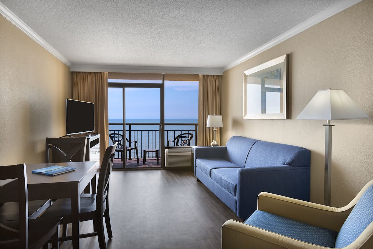 Una habitación de hotel con balcón, sofá, sillón, escritorio y televisión.