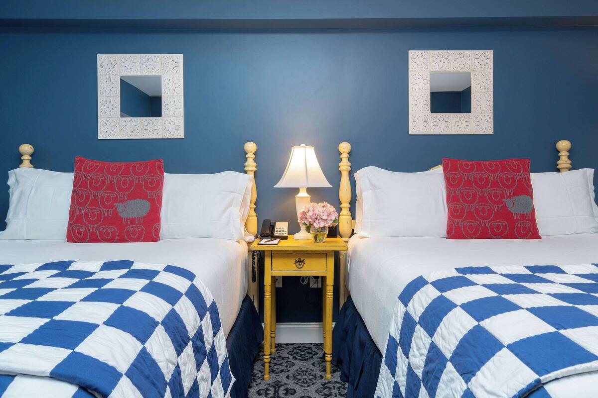 Habitación de hotel con dos camas, cada una con colcha a cuadros azul y blanco, una mesita de noche con lámpara y dos almohadas rojas con estampado de ovejas.