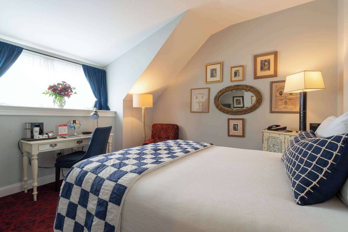 Habitación de hotel con cama, escritorio, silla, lámpara de escritorio, mesita de noche, lámpara y ventana con cortinas.