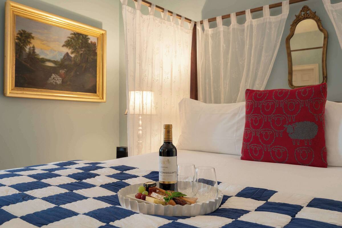 Una cama con colcha de cuadros, una bandeja con comida y una botella de vino, un almohadón rojo con estampado de ovejas y un cuadro en la pared.