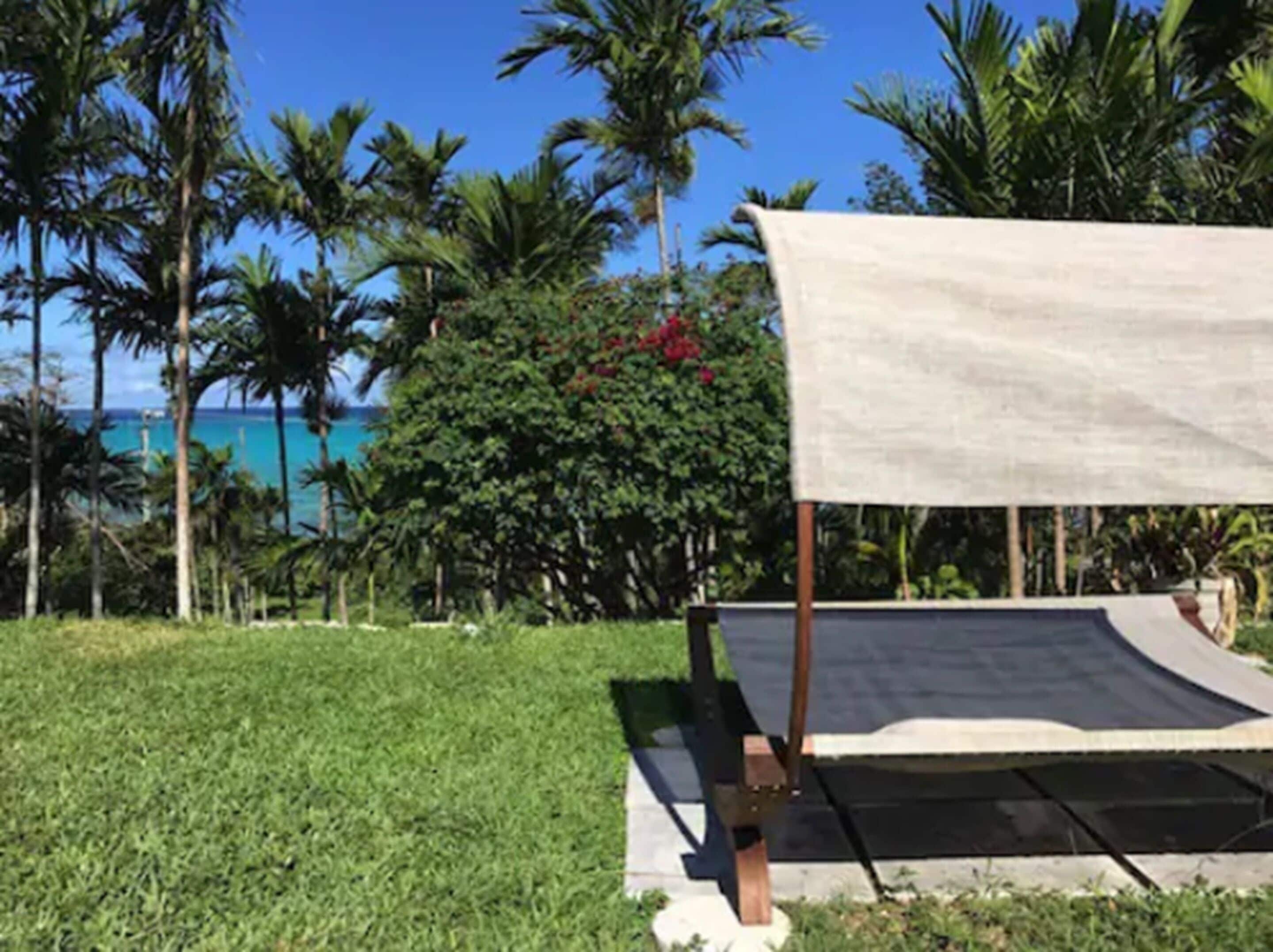 Un área verde con una estructura de toldo blanco, rodeada de palmeras y vista al océano.