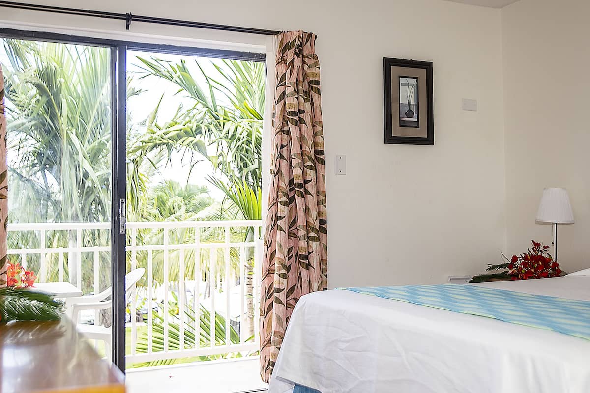 A bedroom with a bed, a nightstand, a lamp, a framed picture on the wall, and a view of greenery through the window.