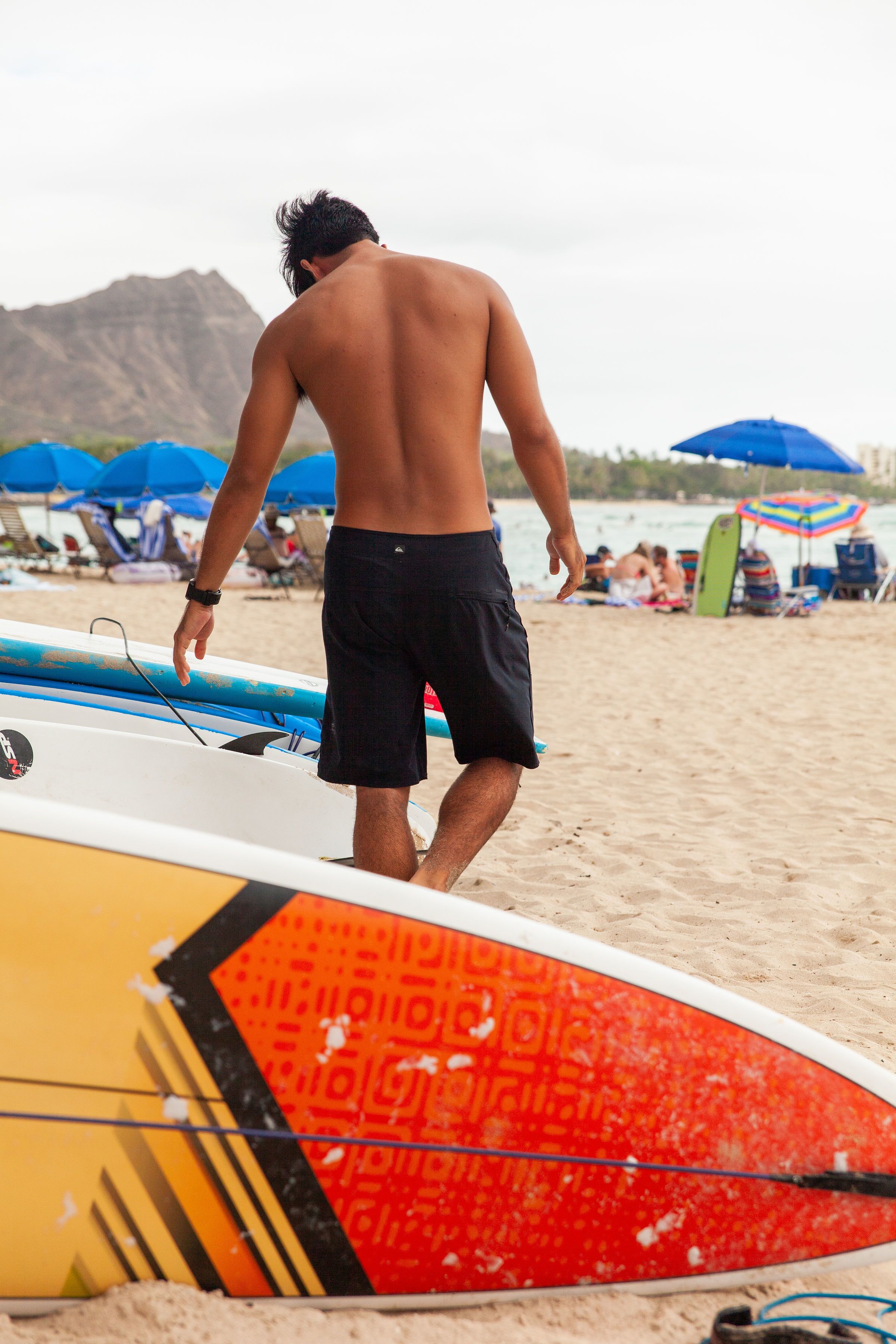 Escena de playa con un hombre caminando hacia el agua, varios tableros de surf y sombrillas de playa al fondo.