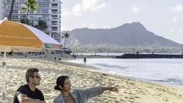 Une scène de plage avec un bâtiment d’hôtel en arrière-plan, un parasol de plage et une montagne au loin.