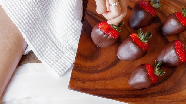 A person in a white robe holding a wooden board with chocolate-covered strawberries. - Vive Hotel Waikiki (Honolulu)