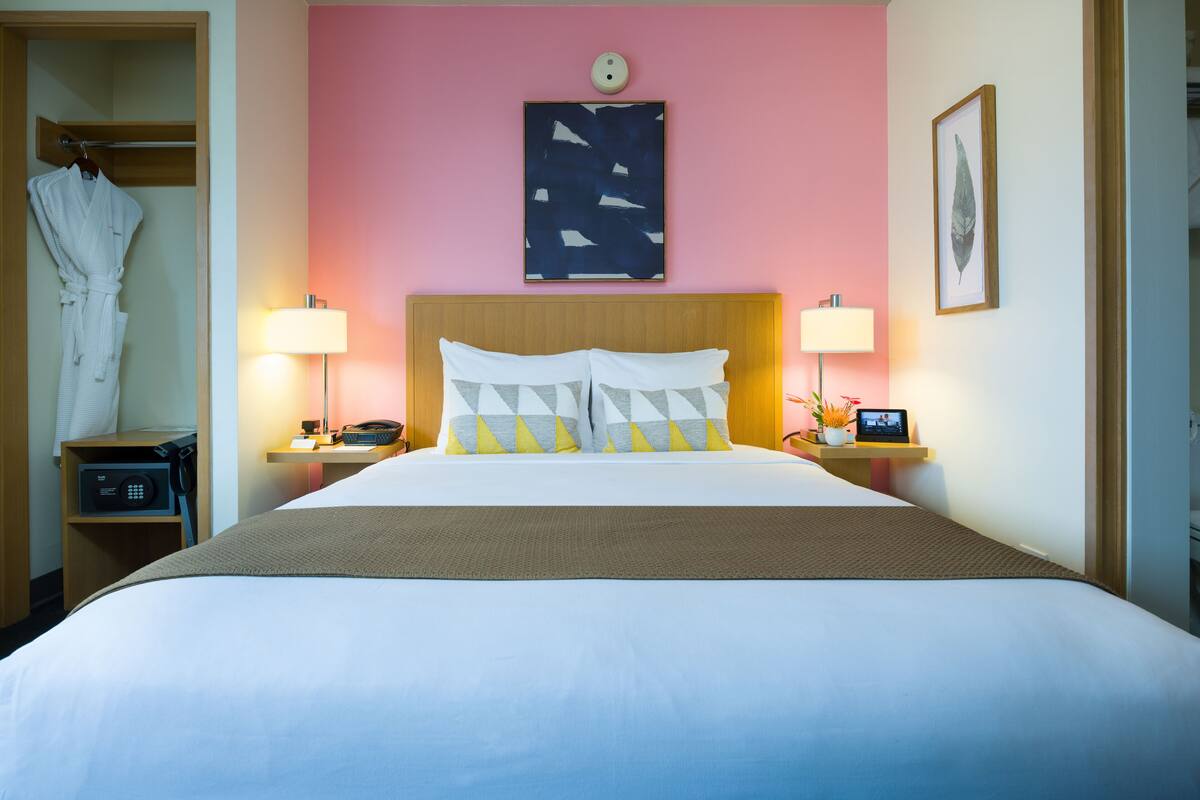 Ein Hotelzimmer mit einem großen Bett, zwei Nachttischen, einem aufgehängten Bademantel, einer Wanduhr und einem gerahmten Bild.