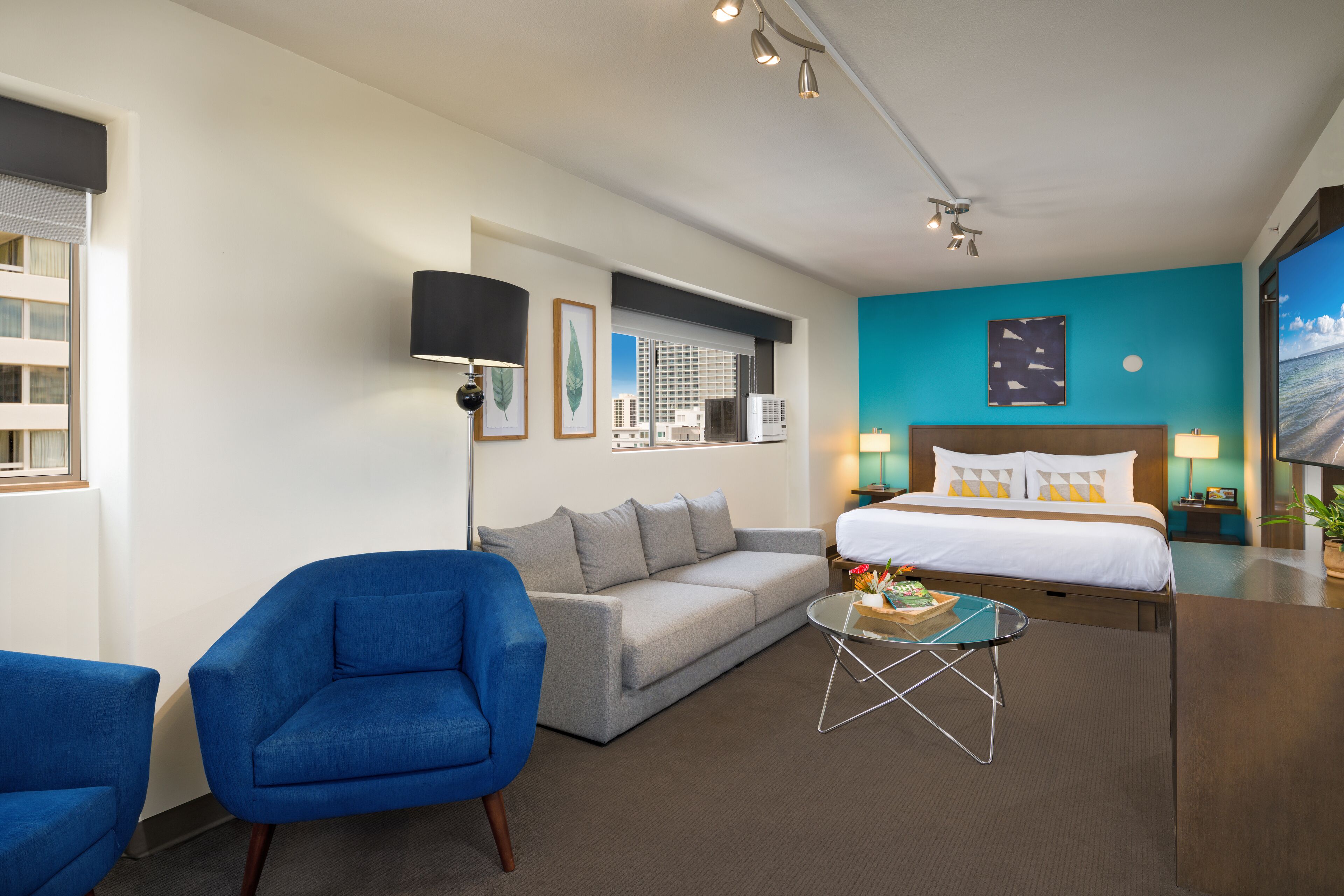 A hotel room with a grey sofa, blue armchair, a small glass coffee table, a bed with white bedding, a teal accent wall, and a large window with a city view.