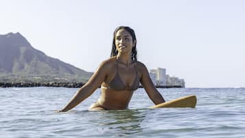 Una persona en traje de baño sobre una tabla de surf en el océano, con una montaña y edificios al fondo.