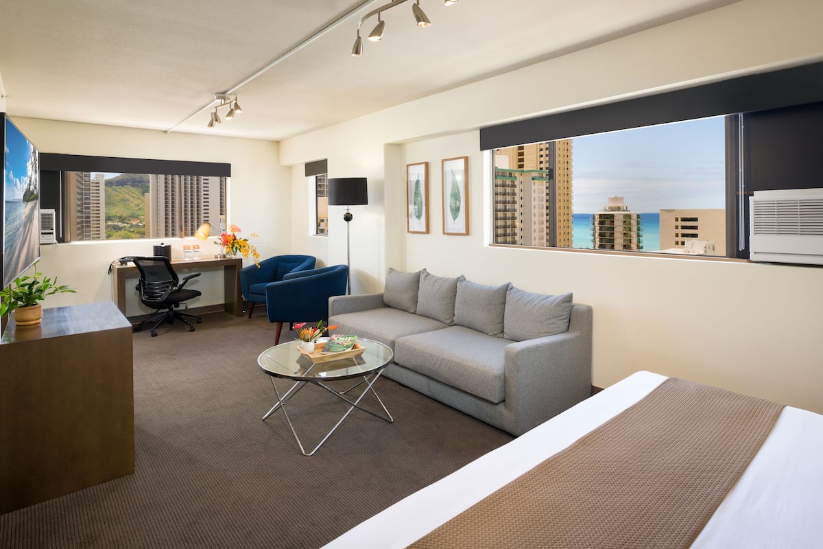 Ein Hotelzimmer mit Bett, Sofa, Schreibtisch mit Computer, Stuhl, Couchtisch und einem großen Fenster mit Blick auf die Stadt.