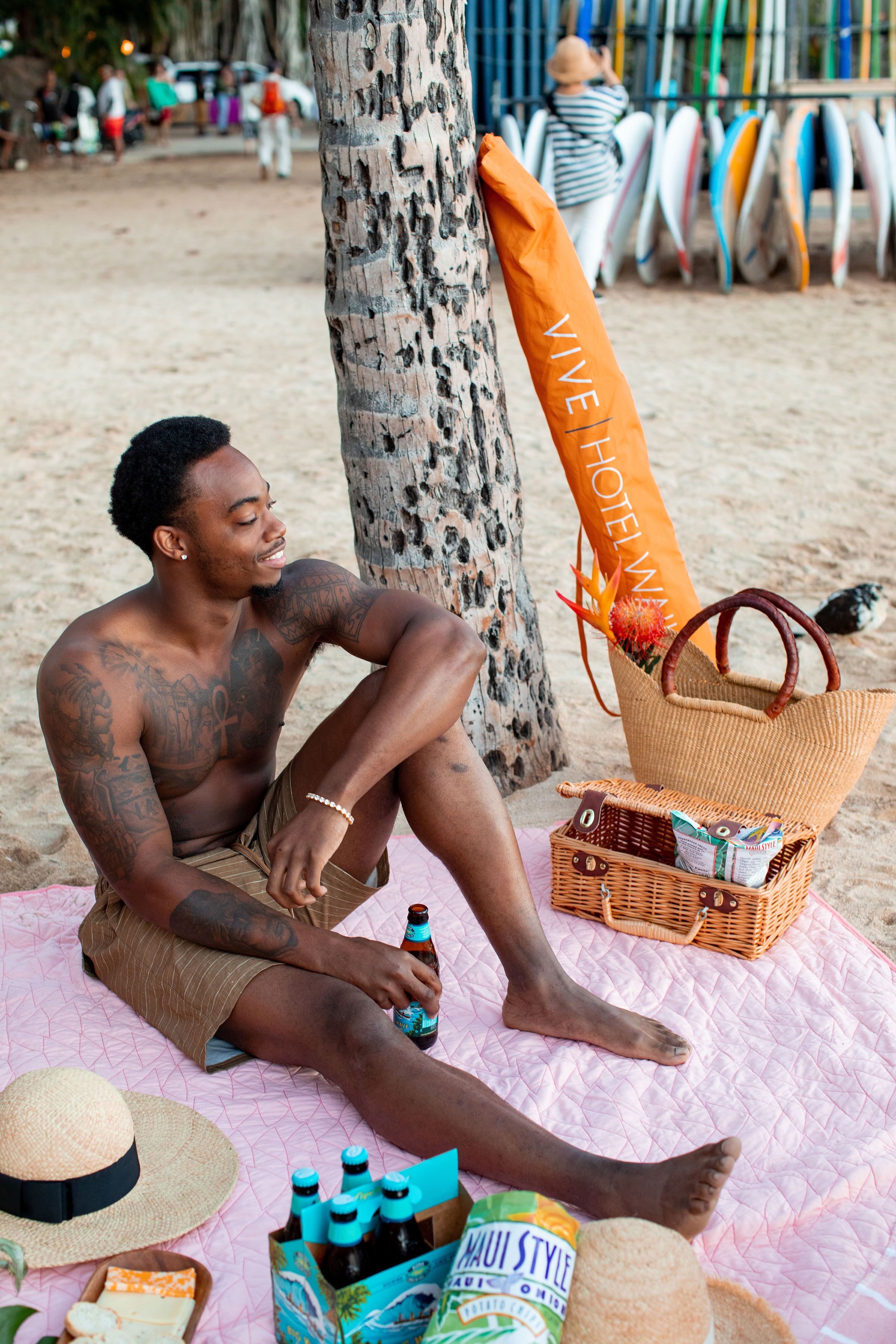 A man sitting on a beach towel with a beach bag, a wicker basket, and bottles.