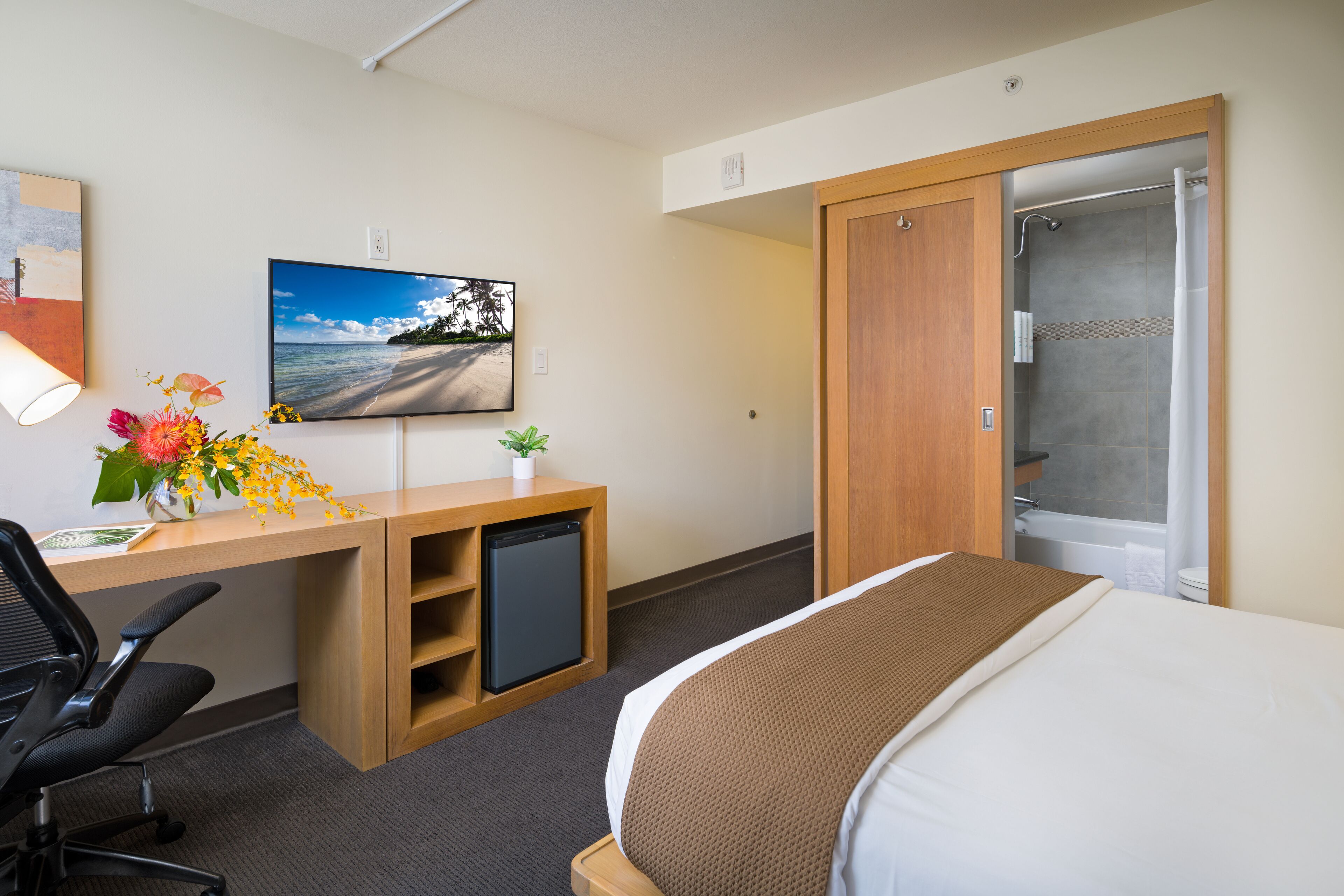 A hotel room with a bed, desk, chair, TV, and a shower area.
