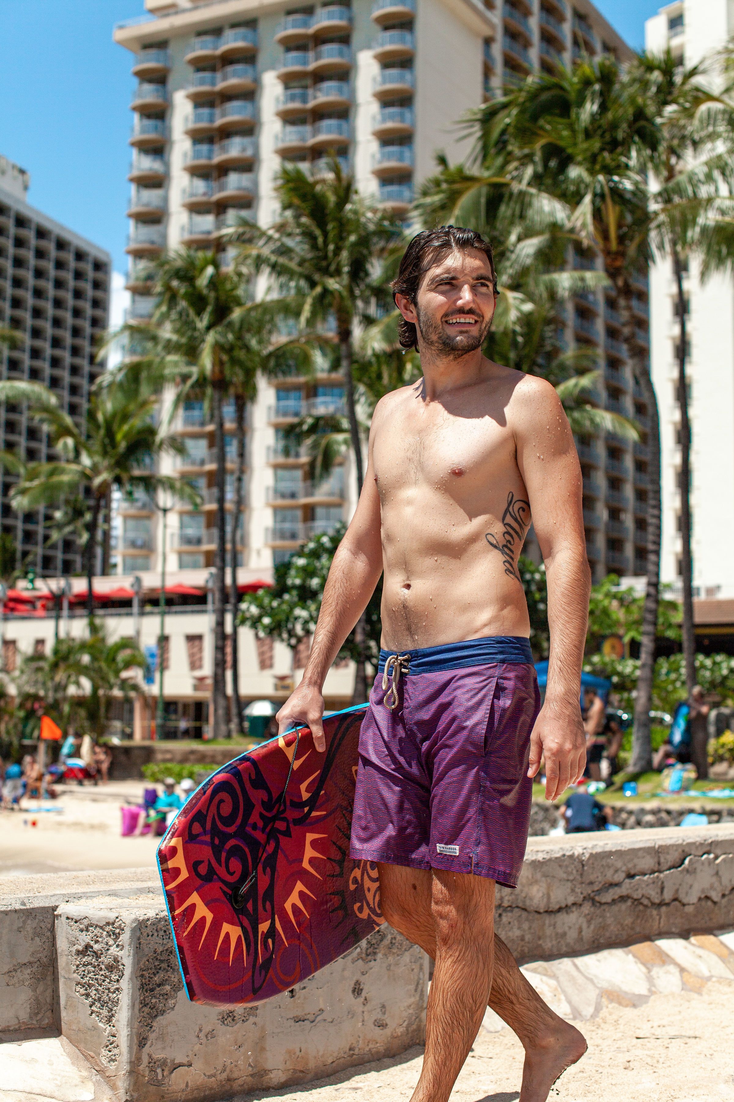 A man walking on a beach with palm trees and a tall building in the background.