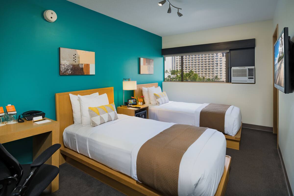 Ein Hotelzimmer mit zwei Betten, einem Schreibtisch mit Stuhl, einem Telefon, einer Lampe und Kunstwerken an den Wänden.