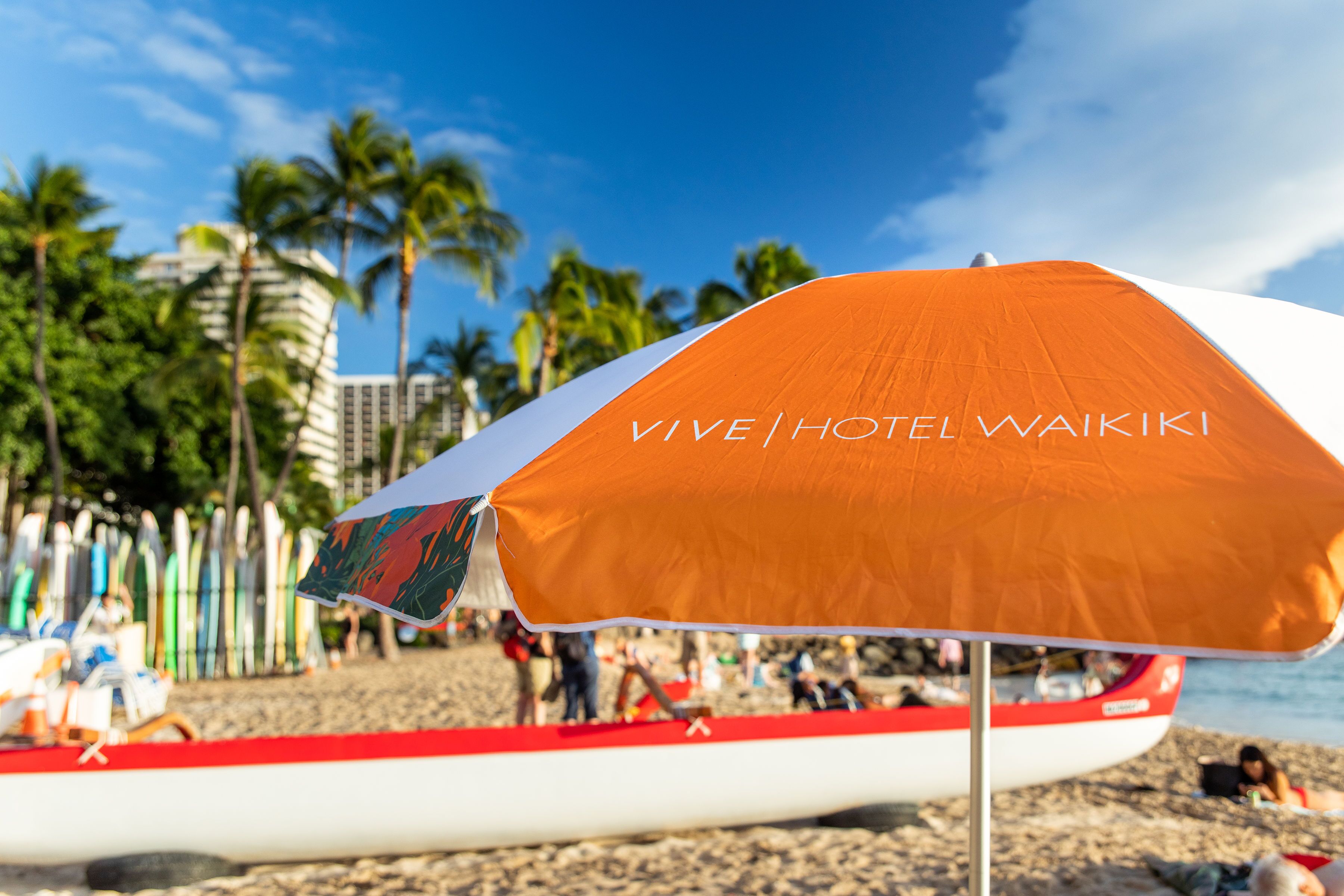 A beach scene with a prominent orange and white umbrella labeled "VIVE HOTEL WAIKI" and a red and white boat in the background.