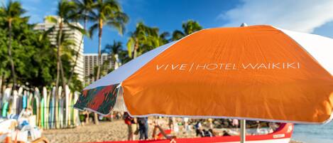 A beach scene with a prominent orange and white umbrella labeled "VIVE HOTEL WAIKI" and a red and white boat in the background.