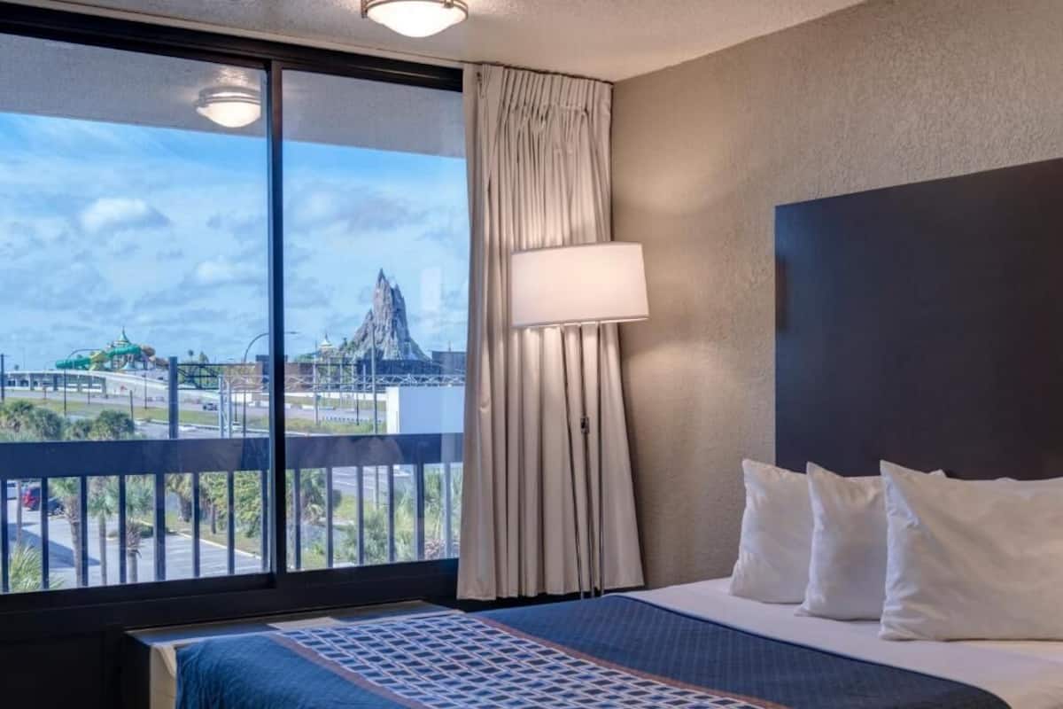 A hotel room with a large window, a bed with white bedding, a lamp, and a view of a mountain.