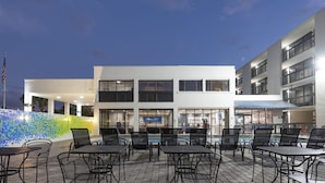 Un área de patio al aire libre con mesas y sillas, una piscina y un edificio de varios pisos con balcones.