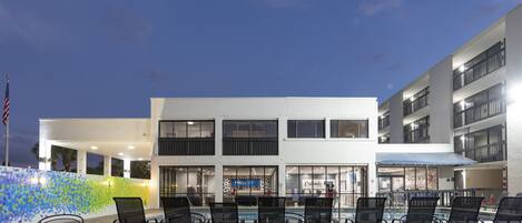Un área de patio al aire libre con mesas y sillas, una piscina y un edificio de varios pisos con balcones.