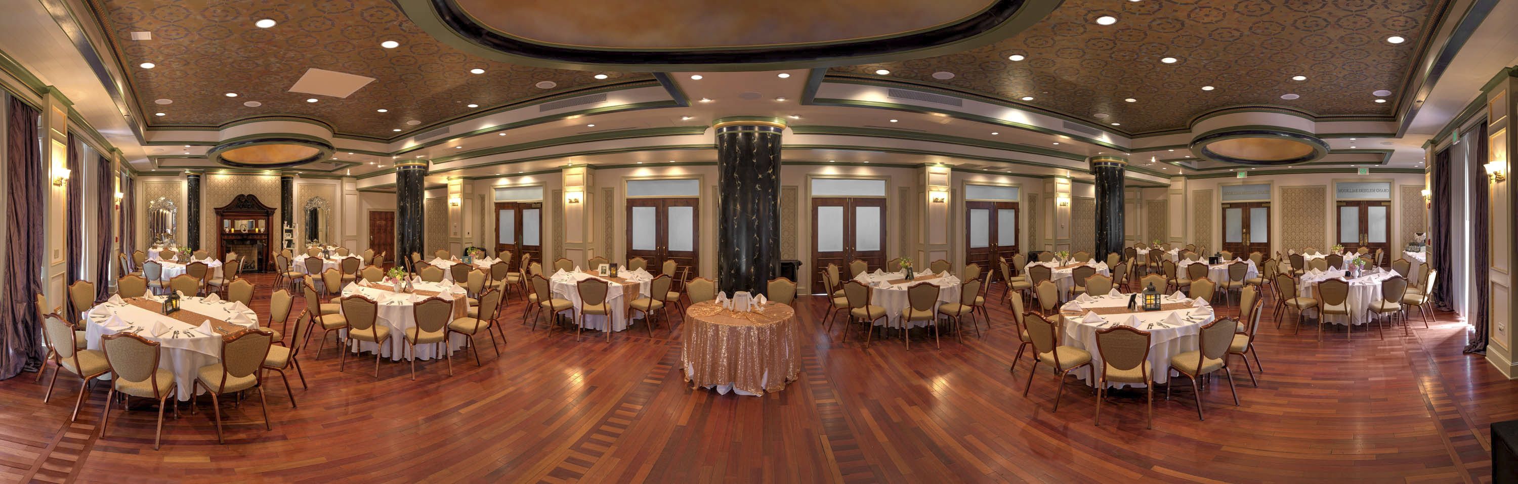 A large banquet hall with multiple round tables, high ceilings with ornate designs, and a polished wooden floor.