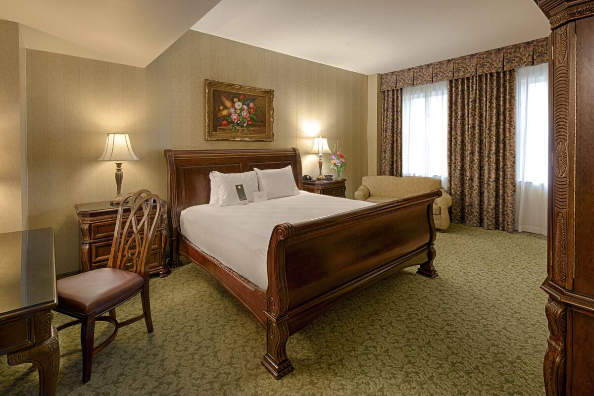 A hotel room with a large wooden bed, a wooden nightstand, a chair, a desk, and a painting on the wall.