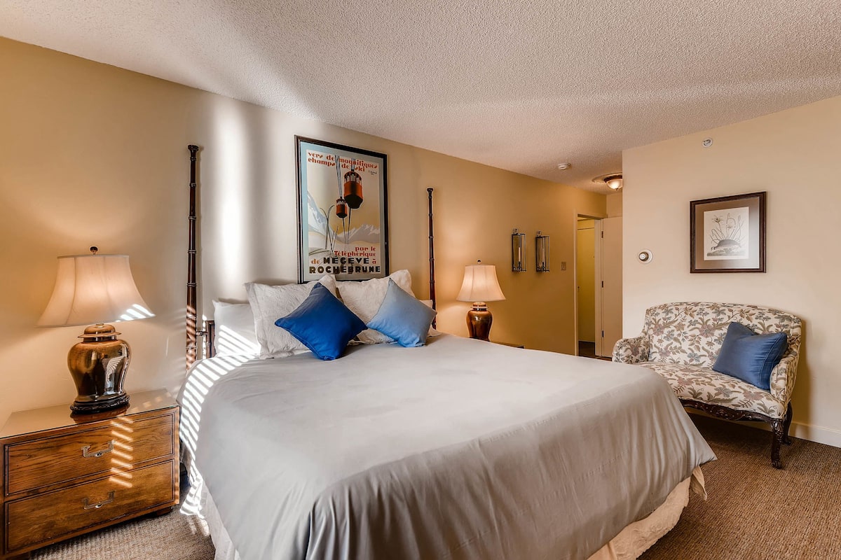 A neatly made bed with a white and blue bedspread, two bedside lamps, a wooden nightstand, a framed poster on the wall, and a patterned armchair.